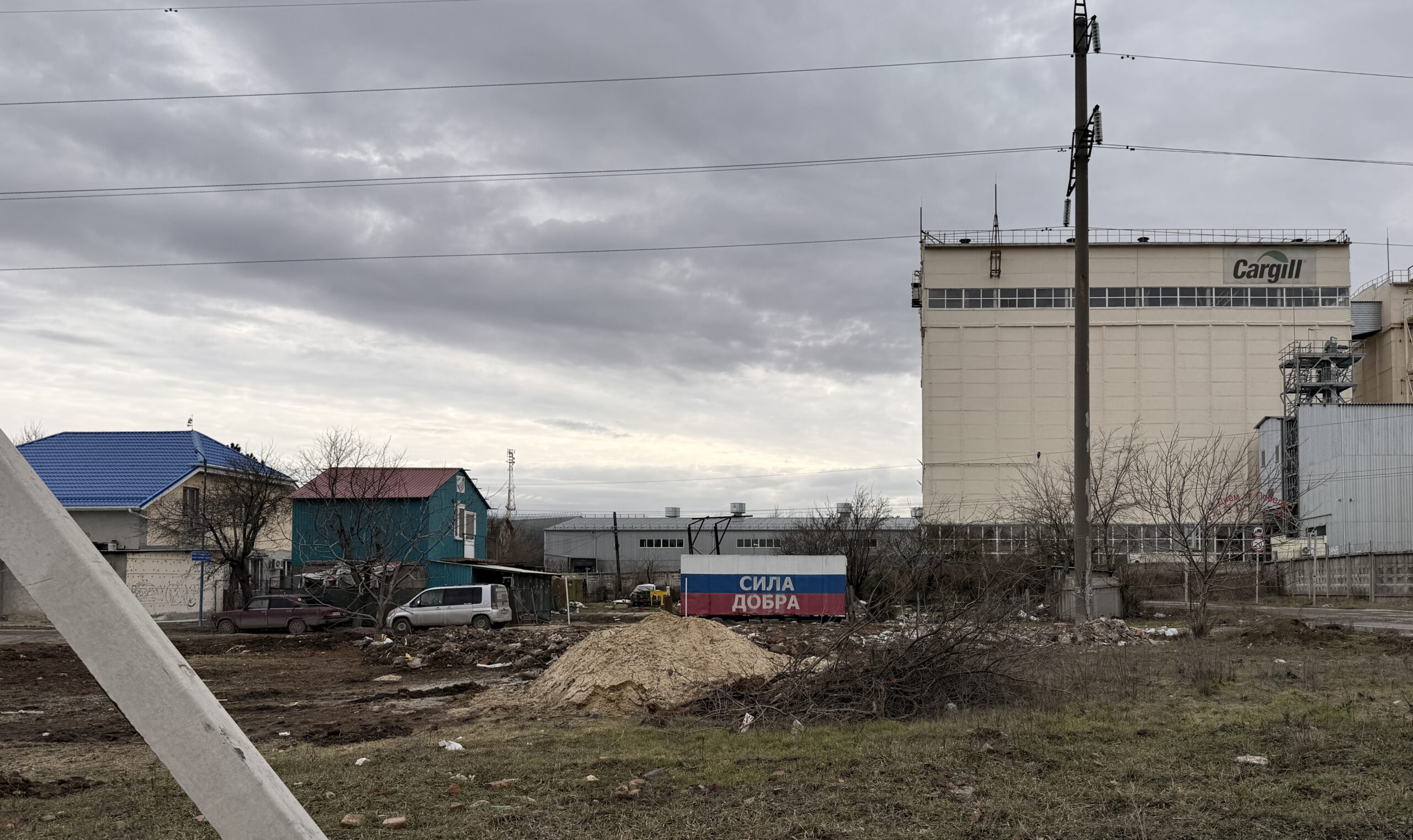 A roadside view near Rostov-on-Don with a factory wall, open ground, houses, wires, and a small structure painted with the words СИЛА ДОБРА