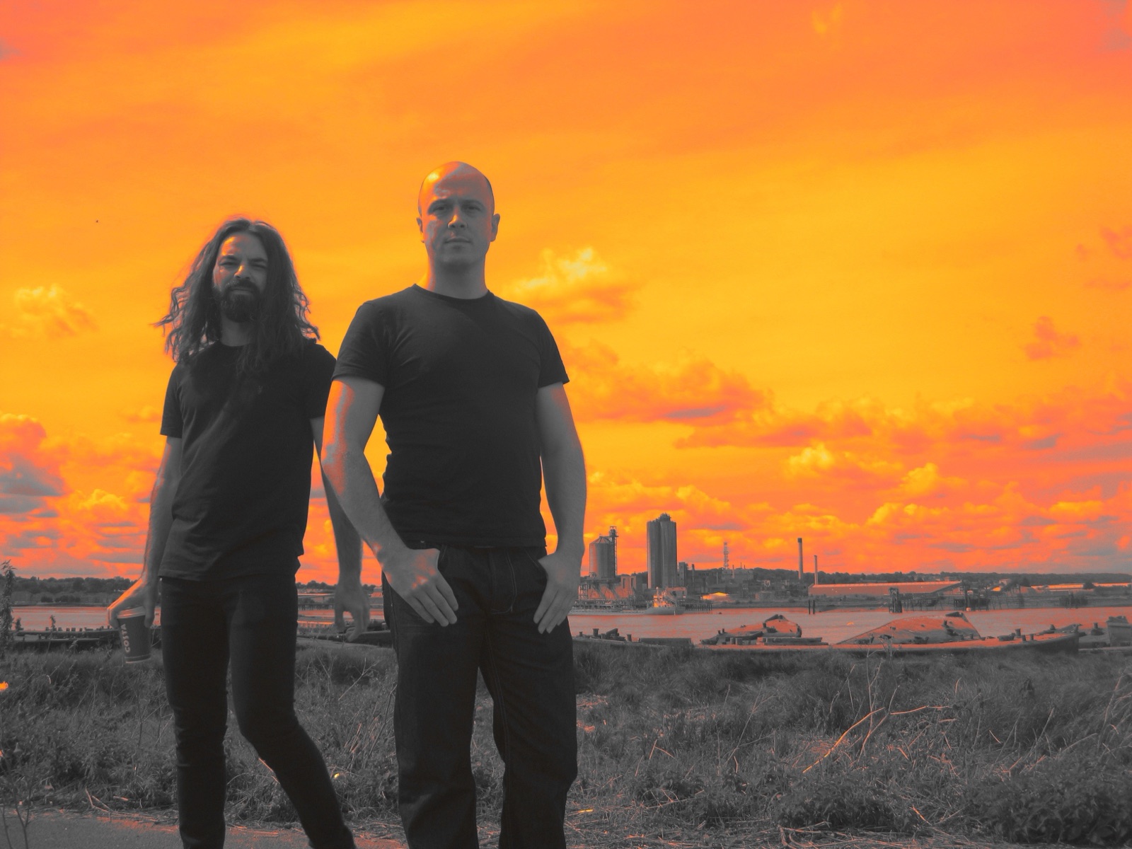 Two musicians in black stand together against an orange industrial waterfront and sky.