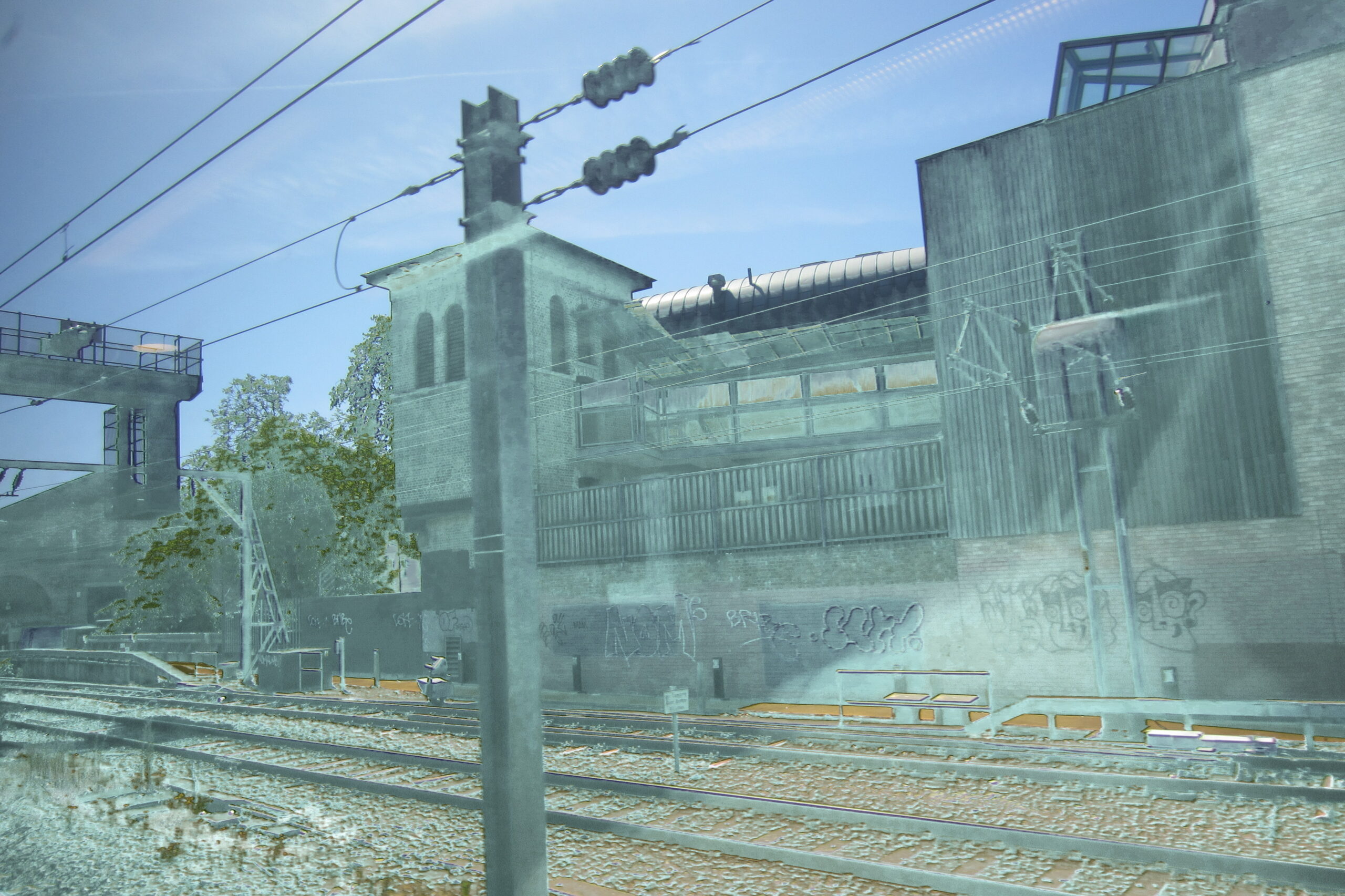 Railway tracks and industrial buildings seen through a train window, divided by a blurred pole.