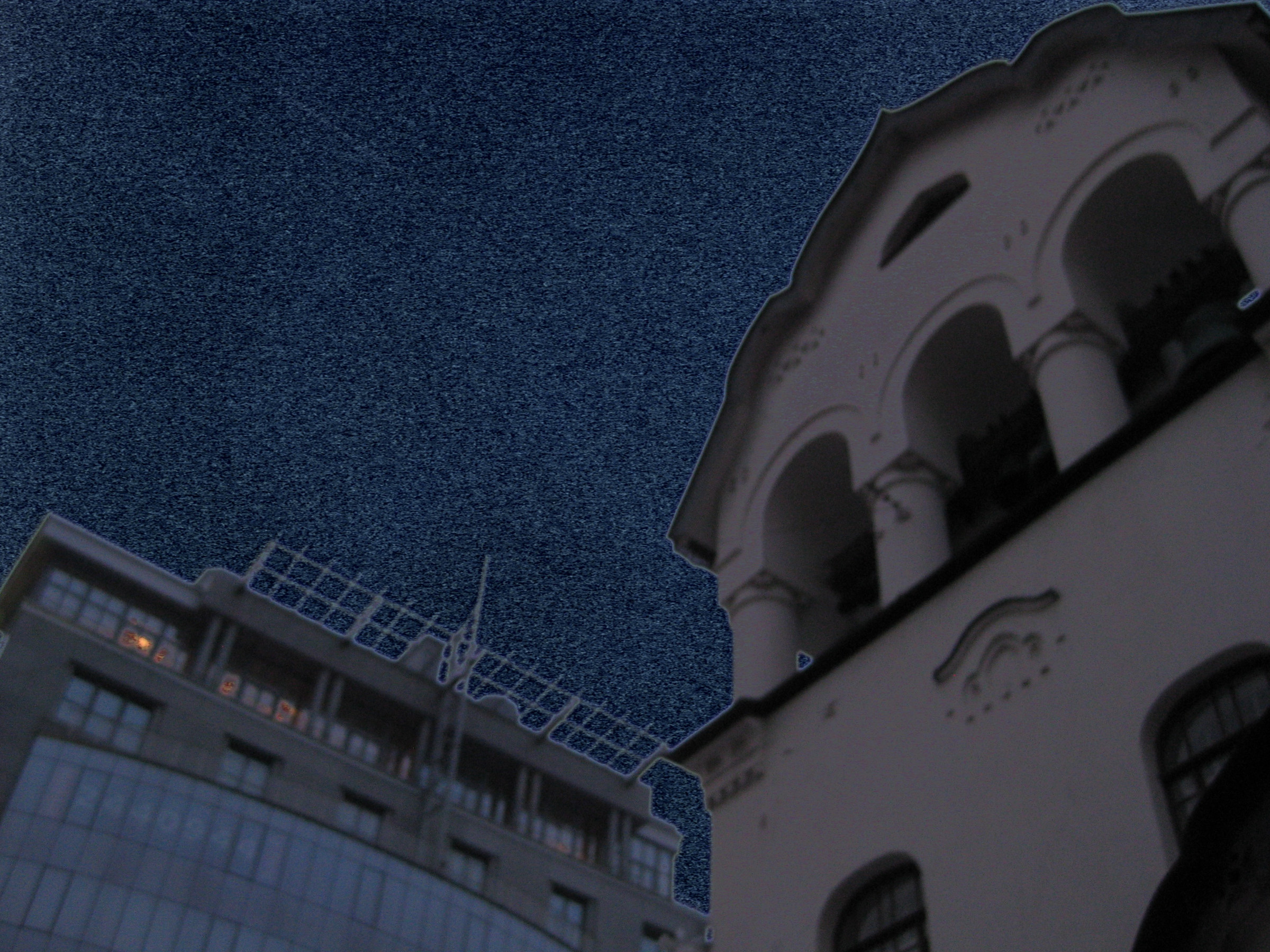 A pale arched facade and a modern building beneath a deep blue grainy sky.