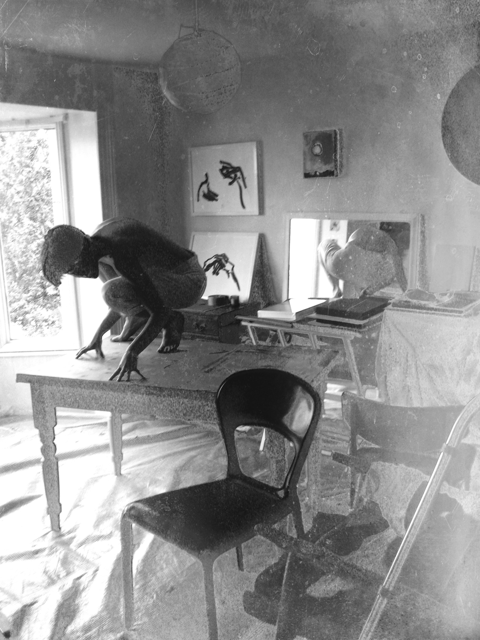 A crouching figure on a table in a room with drawings, a mirror and an open window.