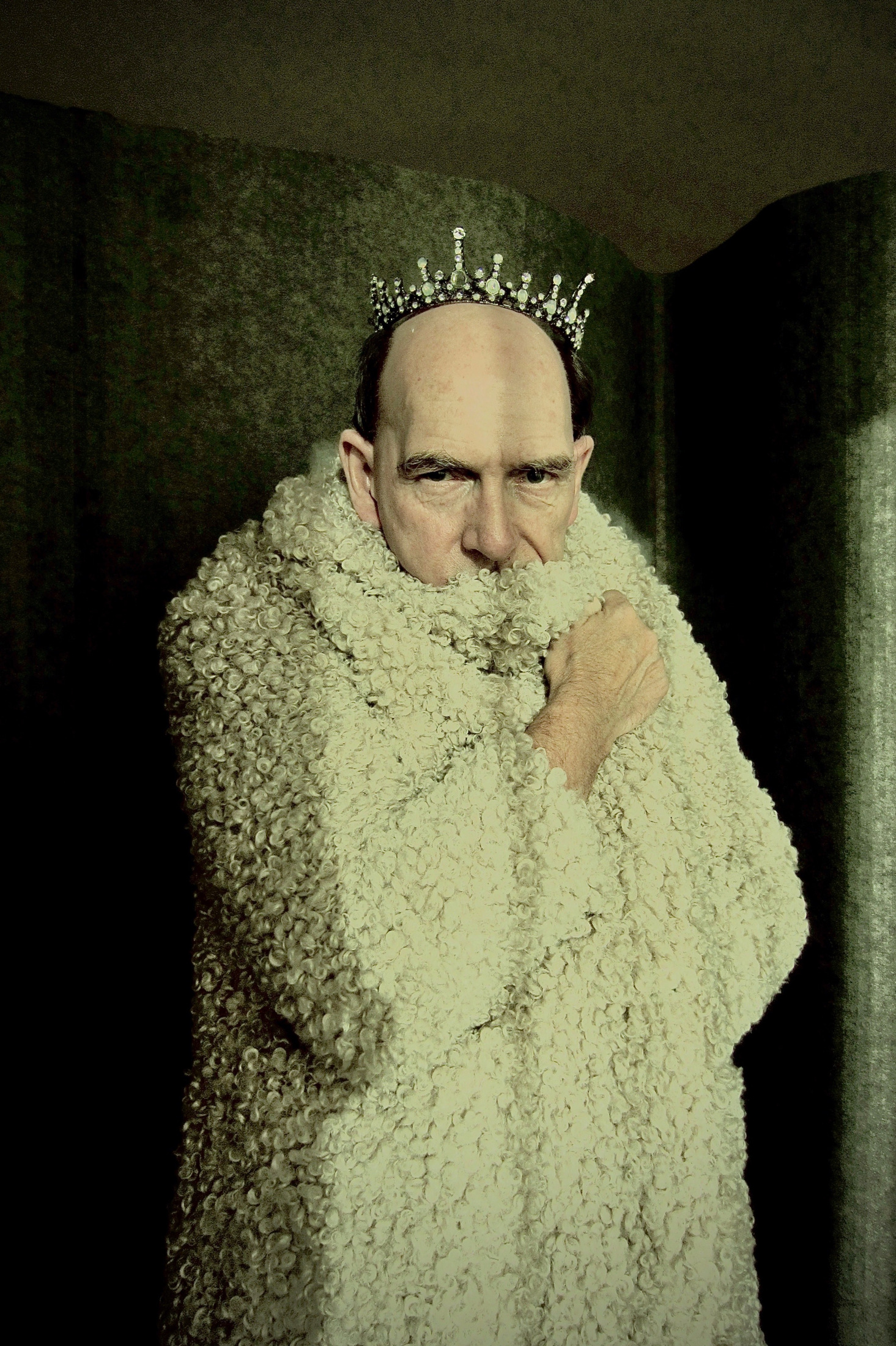 A man wearing a small crown and holding a woolly white wrap around his face.