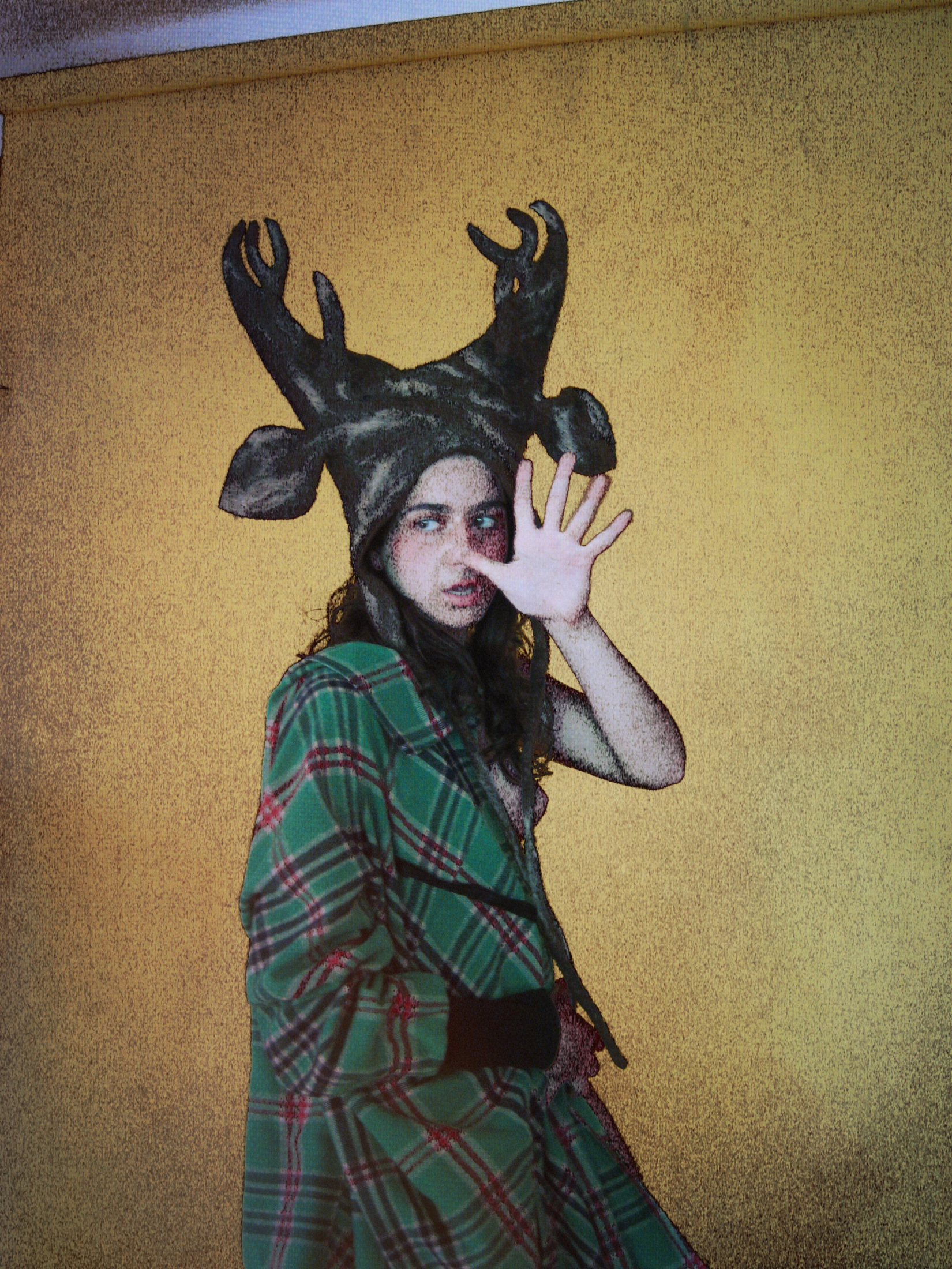 A woman in a green checked shirt wearing an antlered animal head above her face, with one hand raised.